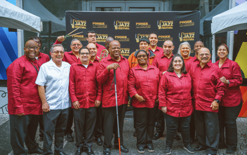 A group of people stands together, smiling in front of a "Jazz Festival" backdrop. They are wearing matching red shirts, except for one person in a white shirt. Tents frame the scene on each side.